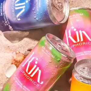 Colorful cans of Kin Euphorics functional beverages with condensation, placed on sand with seashells
