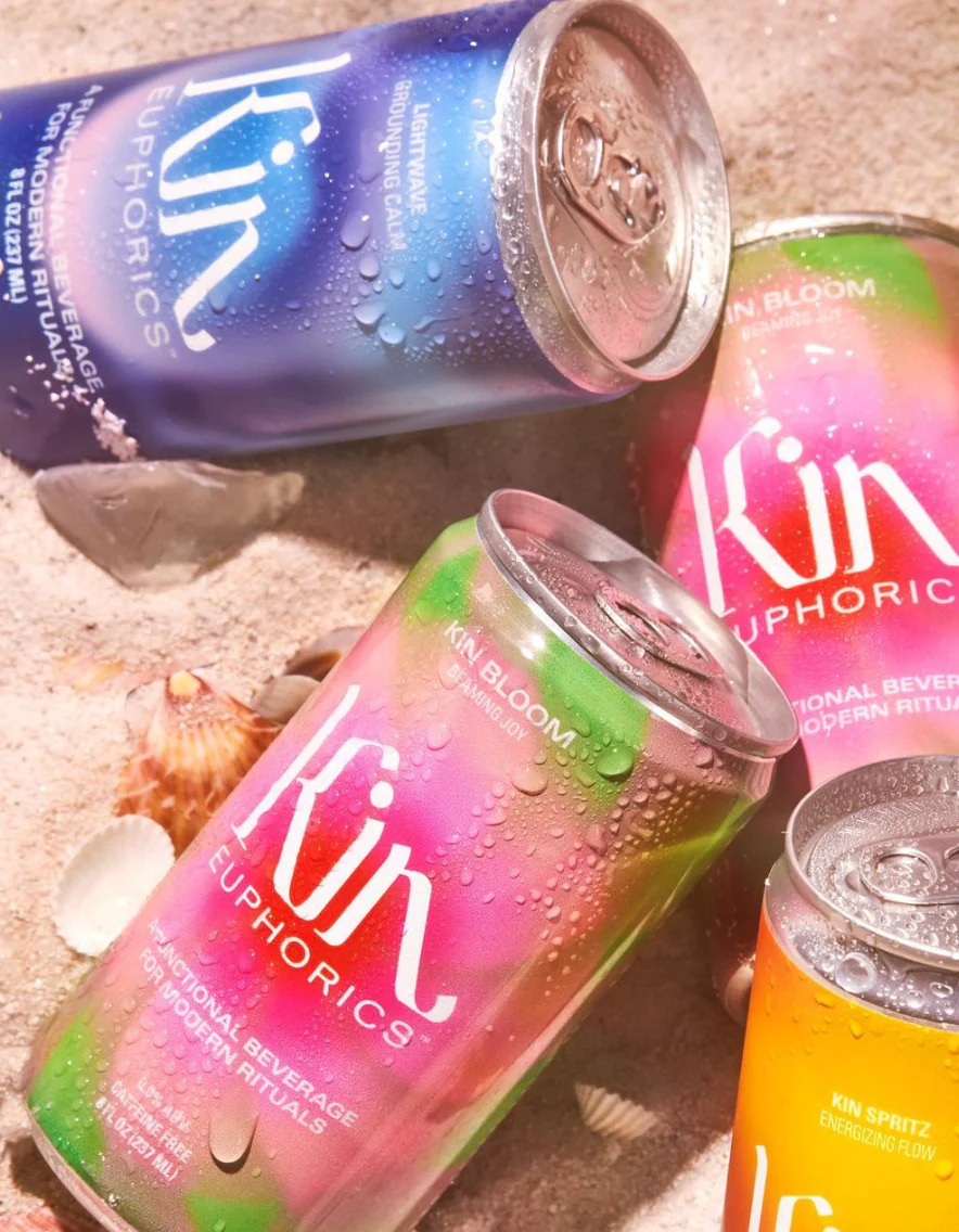 Colorful cans of Kin Euphorics functional beverages with condensation, placed on sand with seashells