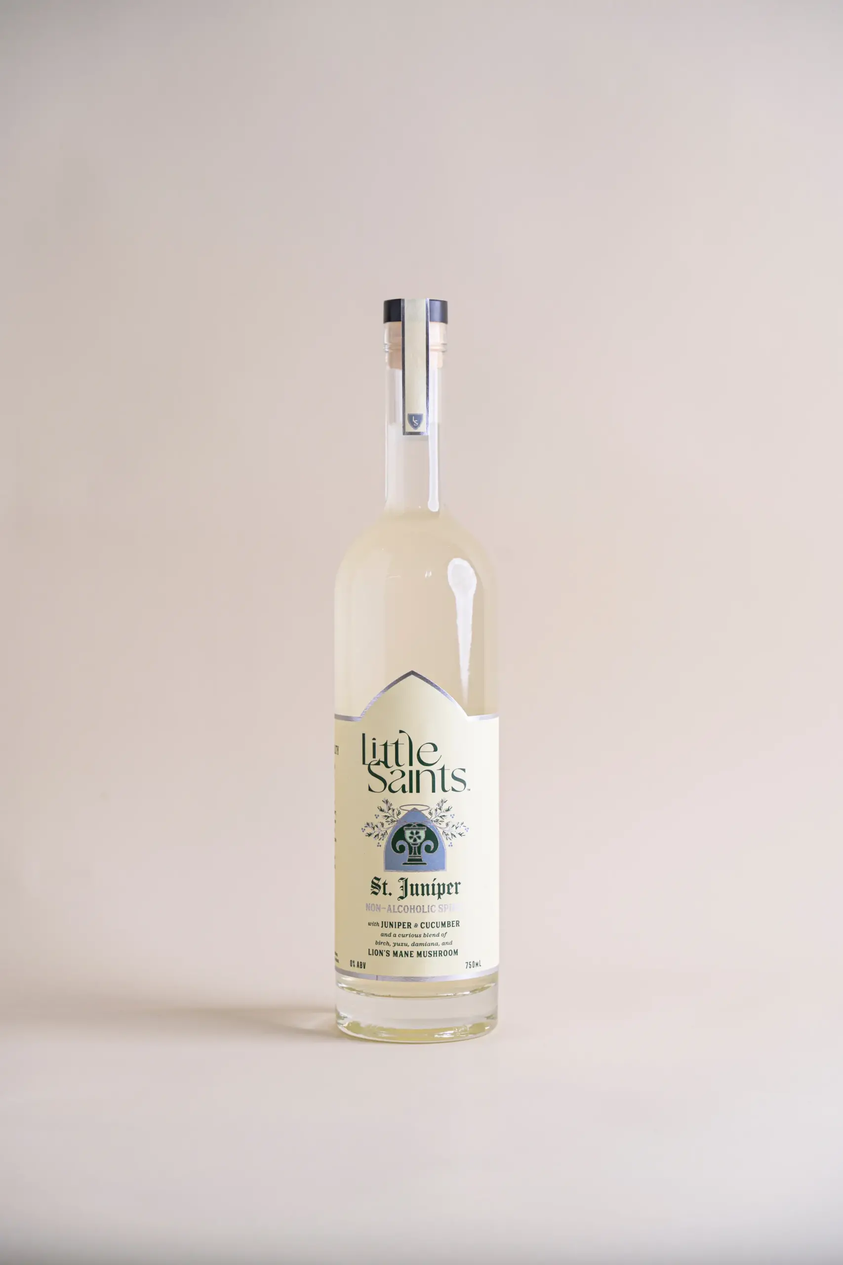 A clear glass bottle of Little Saints St. Juniper non-alcoholic spirit with juniper and cucumber, standing on a neutral beige surface and background