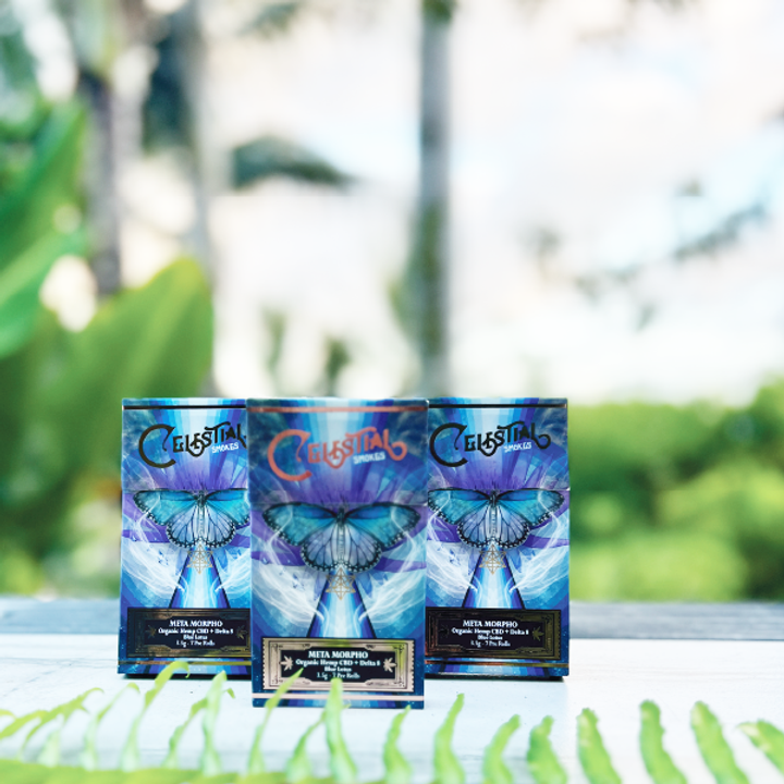 Three boxes of Celestial Smokes Meta Morpho organic hemp CBD pre-rolls displayed outdoors with blurred greenery background.