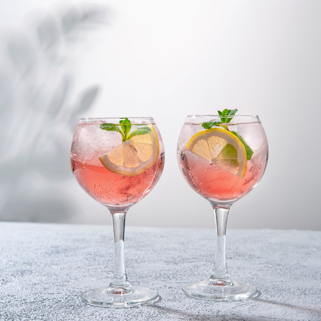 Two glasses of refreshing pink cocktail garnished with lemon slices and mint leaves, served with ice on a light textured surface