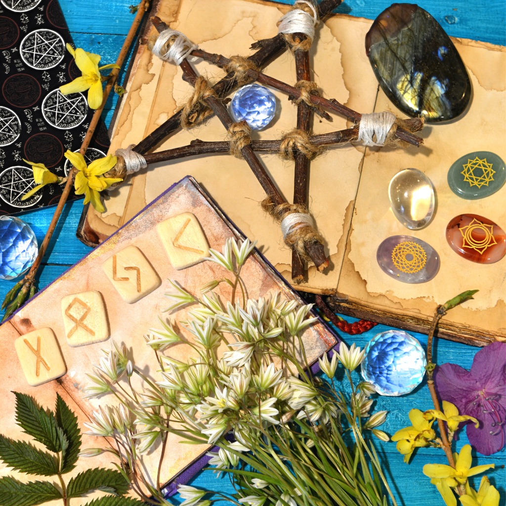 A flat lay arrangement featuring a wooden pentagram, rune stones, chakra crystals, flowers, and an old book on a vibrant blue wooden background, symbolizing spiritual and metaphysical practices