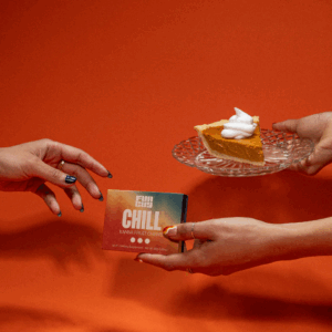 Two hands exchanging a box of Fun Guy Chill Kanna Fruit Chews and a plate with a slice of pumpkin pie topped with whipped cream against an orange background.