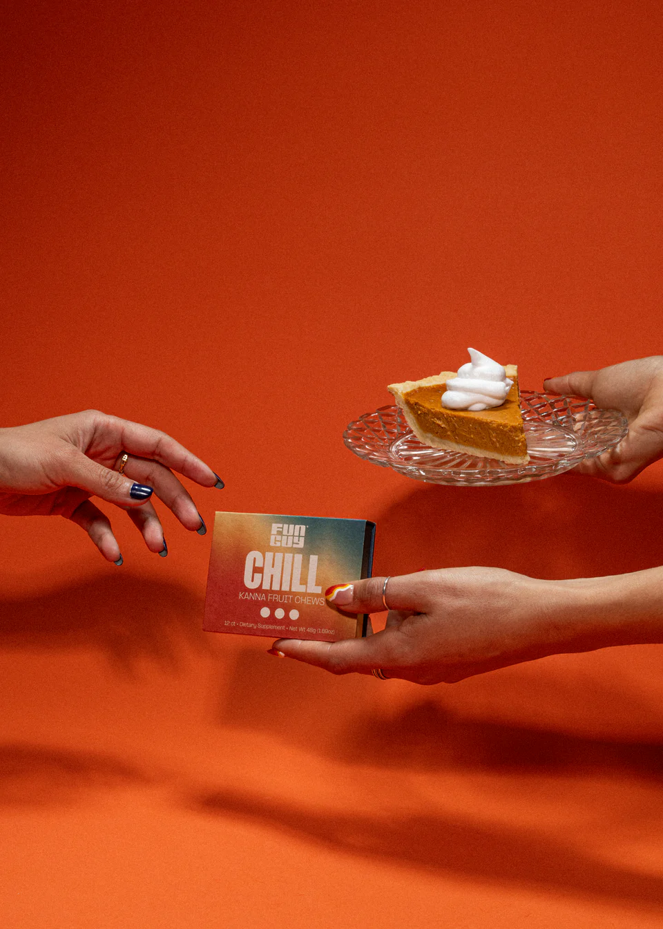Two hands exchanging a box of Fun Guy Chill Kanna Fruit Chews and a plate with a slice of pumpkin pie topped with whipped cream against an orange background.