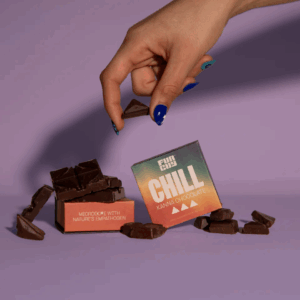 Hand with blue painted nails picking up a piece of dark chocolate next to two colorful boxes labeled 'Chill Kanna Chocolate