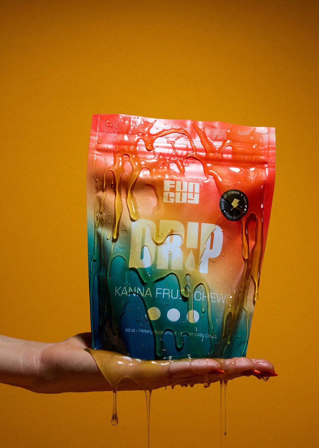 A hand holding a colorful, dripping package of Fun Guy Drip Kanna Fruit Chews against a mustard yellow background.