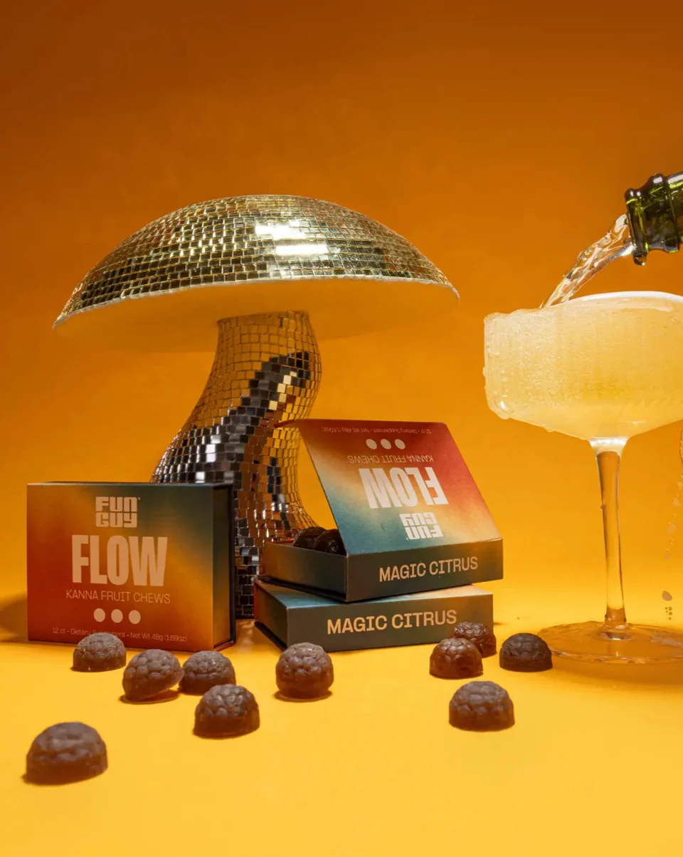 A disco ball mushroom sculpture, Fun Guy Flow Kanna Fruit Chews boxes and candies, and a cocktail being poured against a vibrant yellow-orange background.