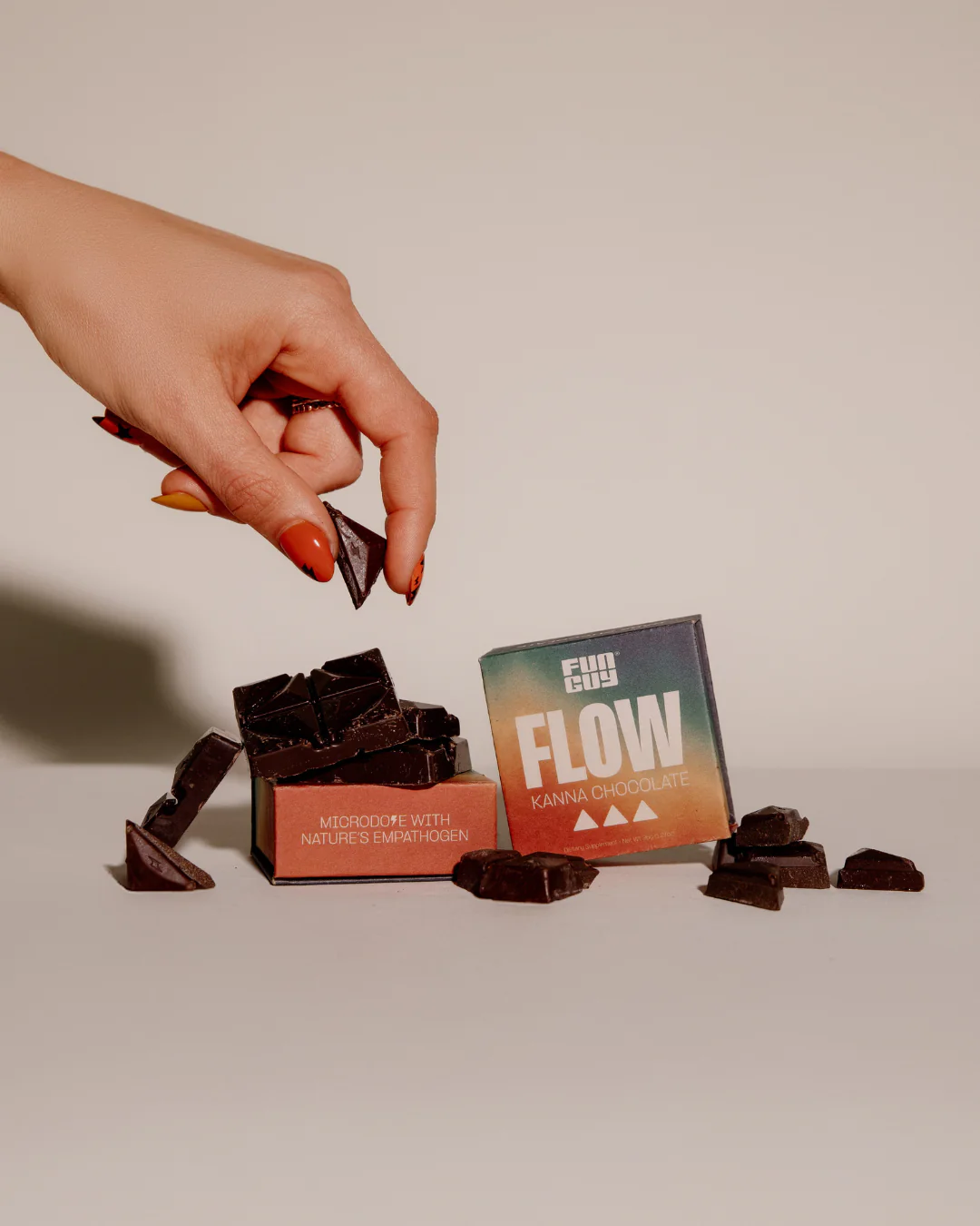 Hand with painted nails picking up a piece of dark chocolate next to two colorful boxes labeled 'Flow Kanna Chocolate
