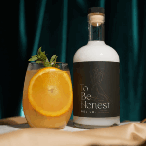 Bottle labeled 'To Be Honest Bev Co.' next to a glass with an orange slice and herb garnish against a dark teal backdrop