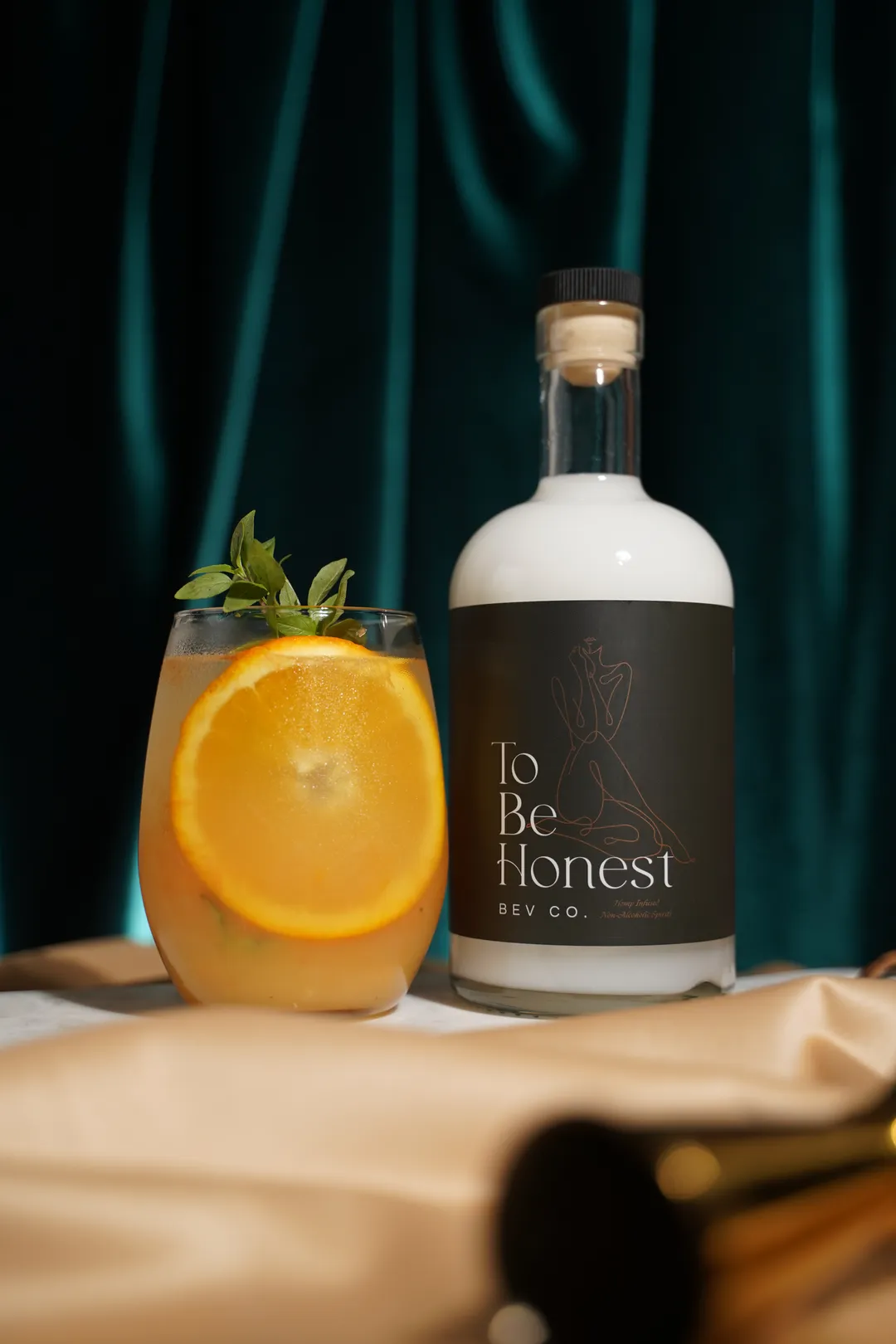 Bottle labeled 'To Be Honest Bev Co.' next to a glass with an orange slice and herb garnish against a dark teal backdrop