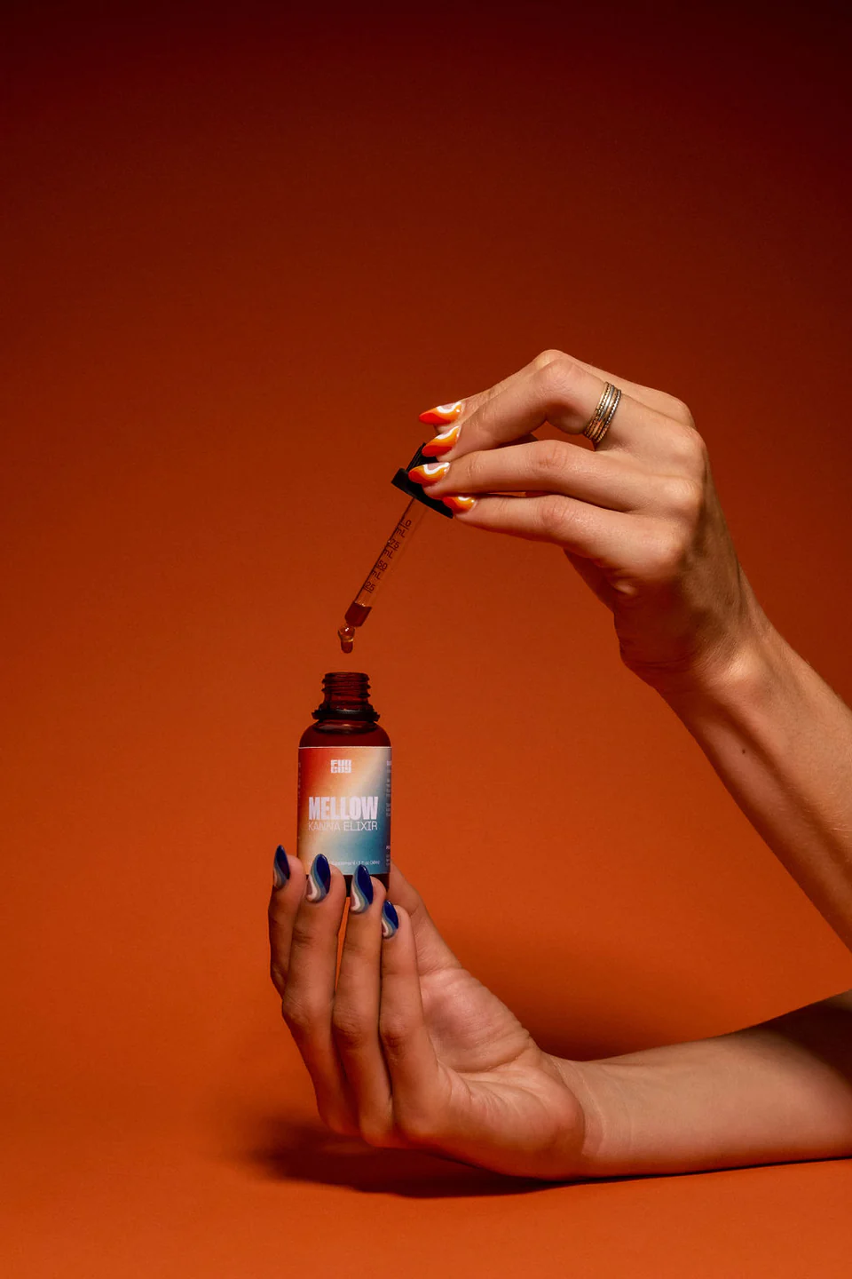 Hands with colorful nails holding a dropper above a small amber bottle labeled 'Mellow.'