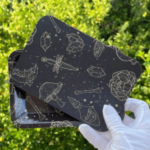 A gloved hand holding a small black rectangular tin with mystical-themed gold illustrations of crystals, bones, mushrooms, and daggers